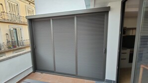 Puerta corredera cerrada con listones horizontales, un balcón con baranda de metal y fachada de edificio con ventanas y contraventanas.