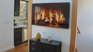 A modern kitchen with dark cabinets, a dishwasher, and a large flat-screen TV mounted on the wall displaying a fireplace image.