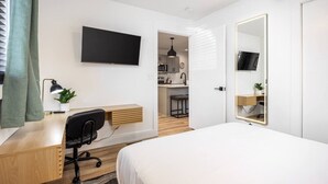 Een moderne hotelkamer met een houten bureau, een zwarte bureaustoel, een flatscreen televisie aan de wand, een grote spiegel en uitzicht op de keuken.