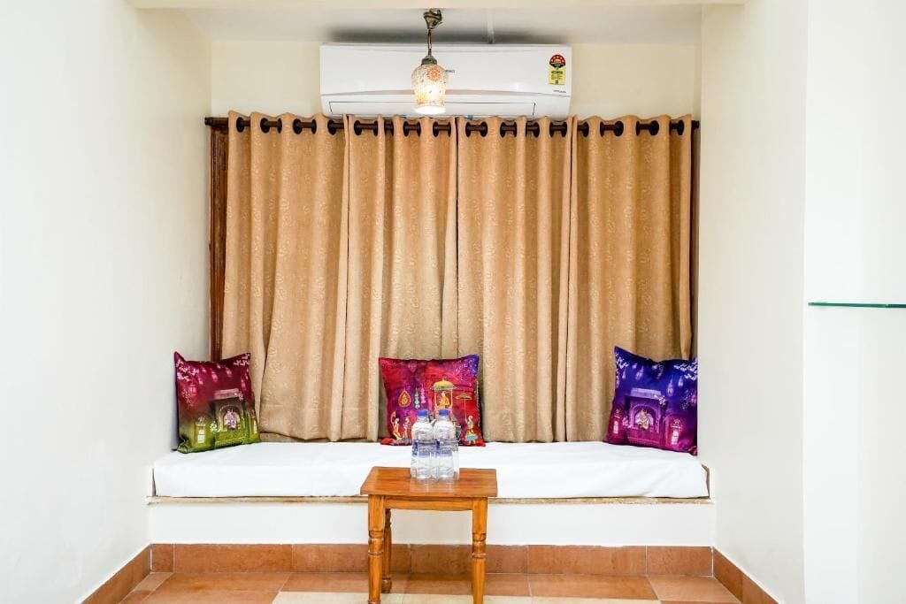 A room with a white sofa, two colorful pillows, a wooden table with water bottles, and a beige curtain.