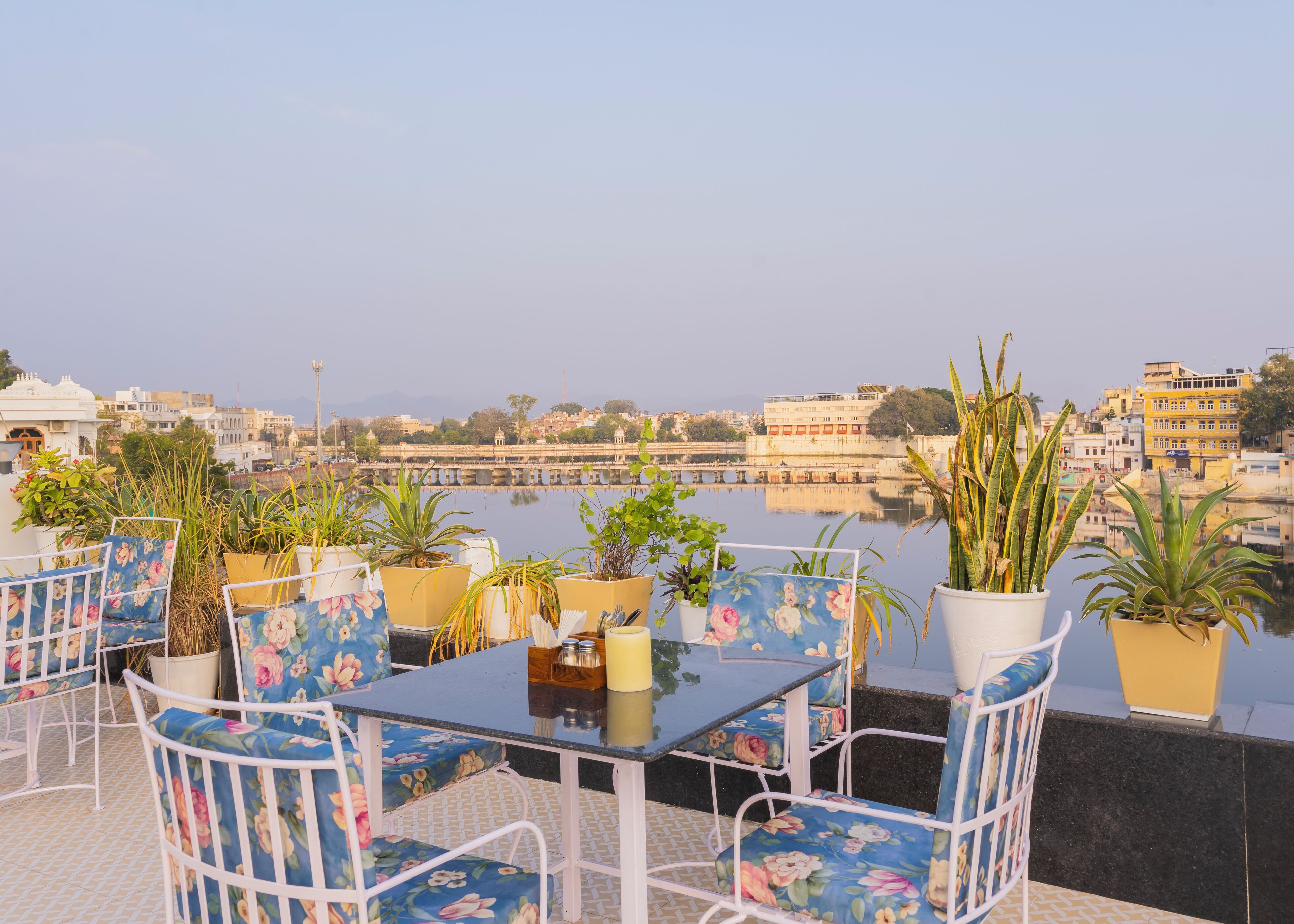 A terrace with a table and chairs, potted plants, and a view of a river and buildings.