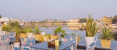 A terrace with a table and chairs, potted plants, and a view of a river and buildings.