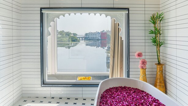 A bathroom with a bathtub filled with rose petals, a window with a view of a river and buildings, and two wooden vases with flowers. - Natural Lake View Hotel (Udaipur)