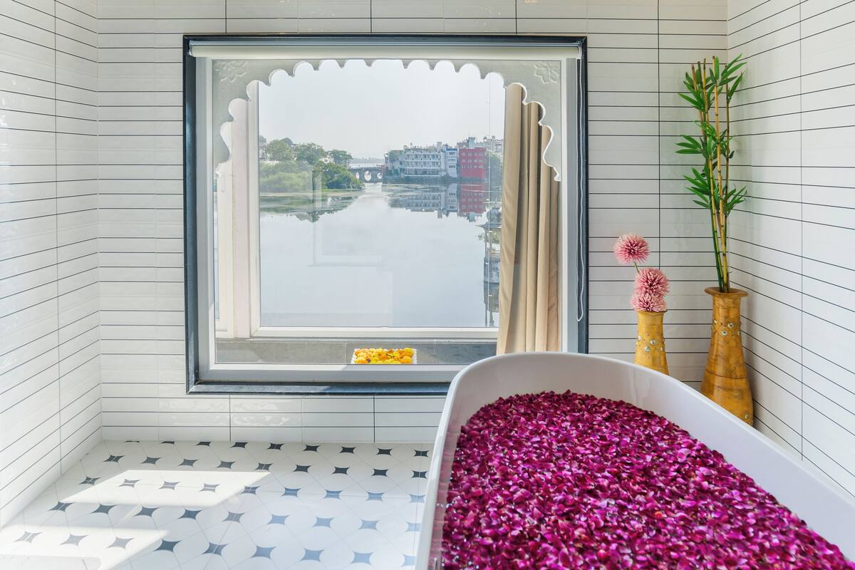 A bathroom with a bathtub filled with rose petals, a window with a view of a river and buildings, and two wooden vases with flowers.