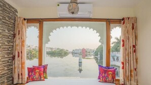 A room with a bed, a large window with a decorative arch, and a view of a water body and buildings. - Natural Lake View Hotel (Udaipur)
