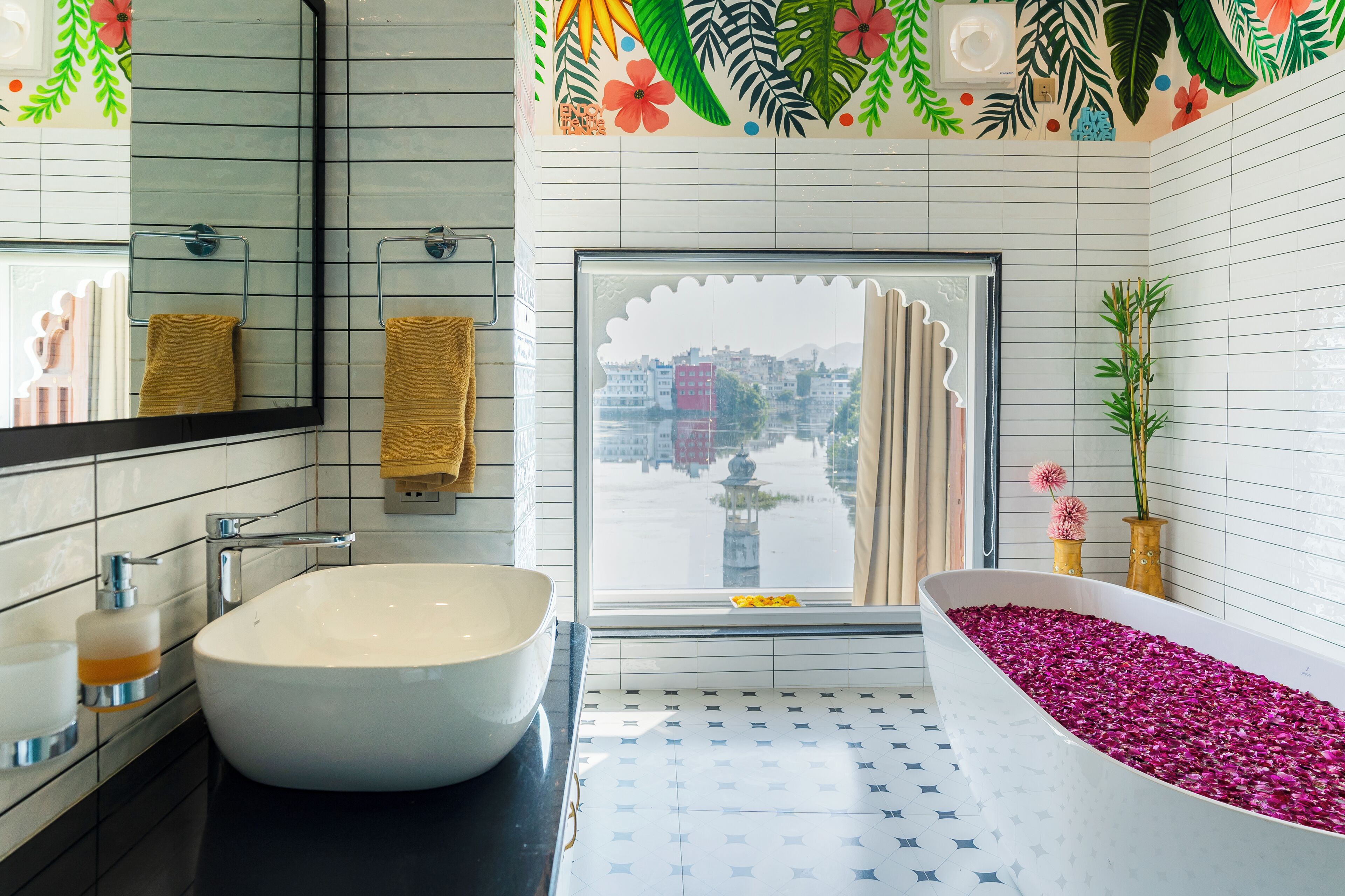Une salle de bain moderne avec un grand lavabo, une baignoire remplie de pétales de roses, une fenêtre donnant sur des immeubles et un miroir décoratif.