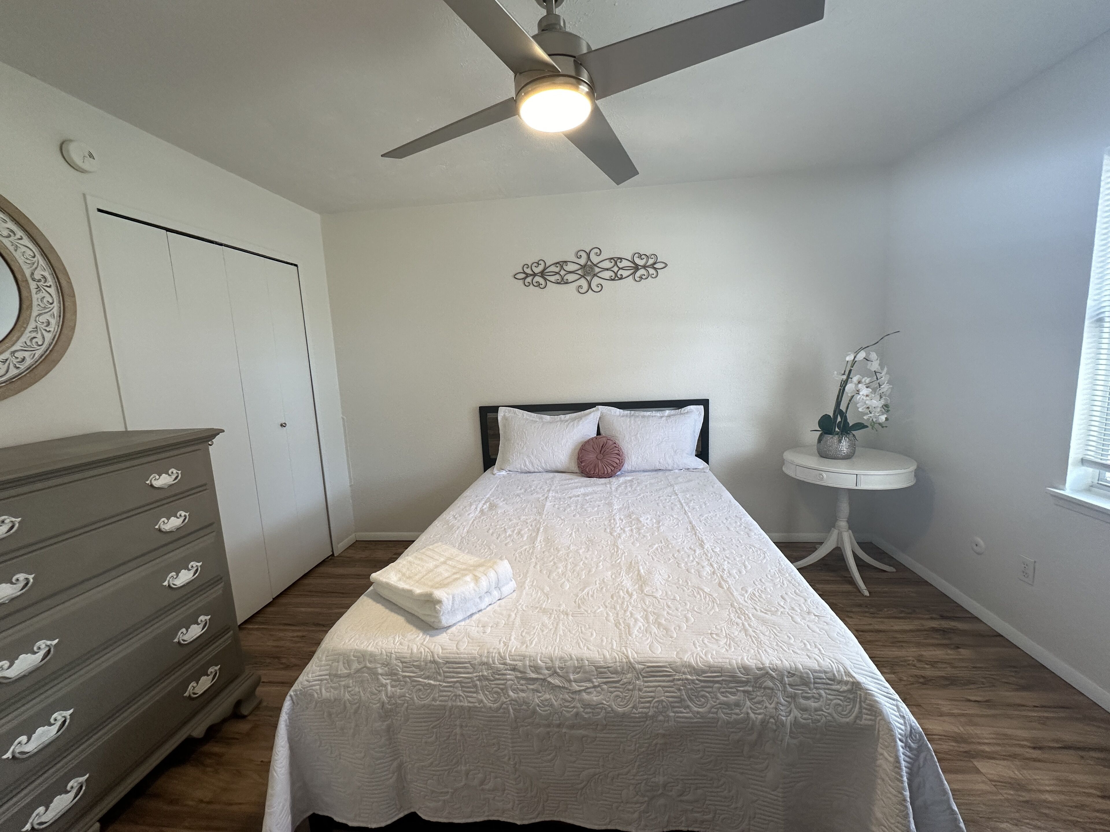 Ein Schlafzimmer mit Bett, Schminktisch, Deckenventilator, Wanddekoration und einem kleinen Tisch mit Pflanze.