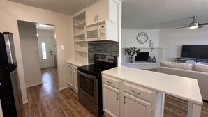 A modern kitchen with white cabinetry, a black stove, and a microwave. There is a white countertop and a ceiling fan in the living area.