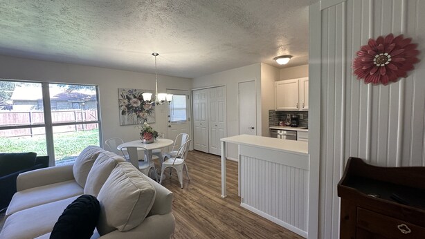 A spacious living room with a white sofa and a dining area with a round table. A kitchen with white cabinets and a red flower decoration is visible.
