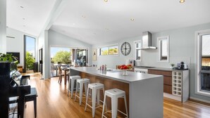 A modern kitchen with a large island, wooden flooring, and a piano in the adjacent room. - Jervis Bay Sanctuary by Experience Jervis Bay (Vincentia)