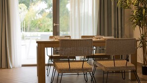 Comedor con mesa de madera y sillas de mimbre, una planta en maceta y vistas a través de un ventanal.