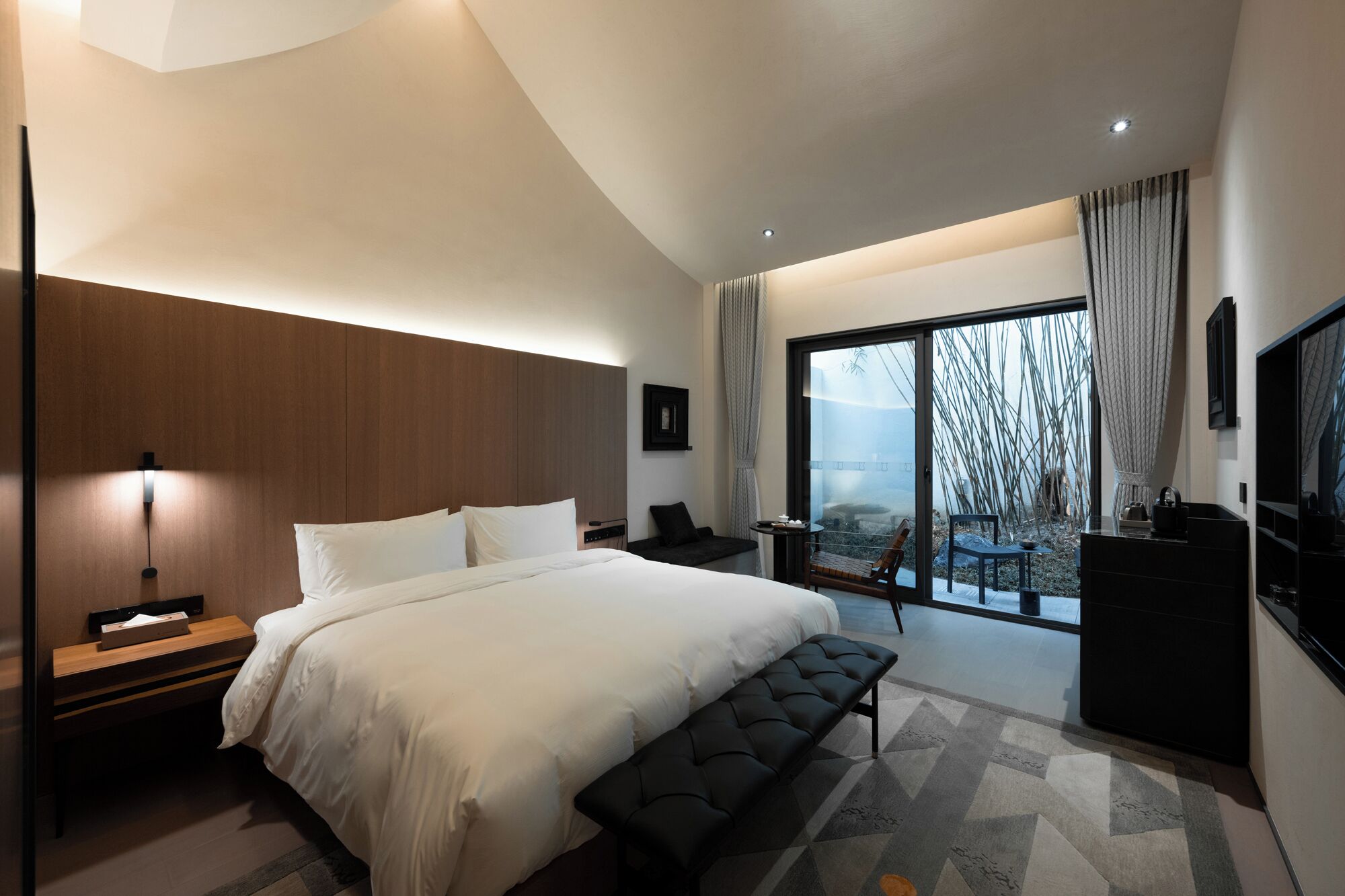 A modern hotel room with a large bed, a bench, a desk, and a view of the outdoors.