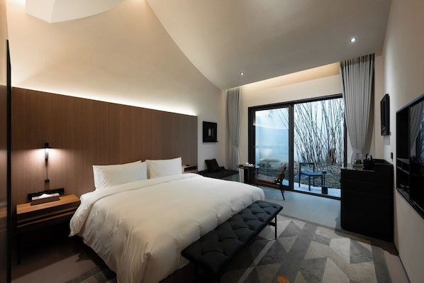 A modern hotel room with a large bed, a bench, a desk, and a view of the outdoors.
