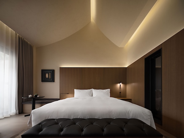 Quarto de hotel moderno com uma cama grande, uma mesinha de cabeceira, uma luminária e vista para o exterior.