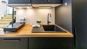 A modern kitchen with a wooden countertop, a black sink, and a stainless steel faucet. There is a small island with a glass top and a black appliance. The kitchen has dark cabinets and a light-colored backsplash.