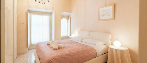 Un dormitorio con cama, mesita de noche con lámpara, candelabro y dos ventanas.