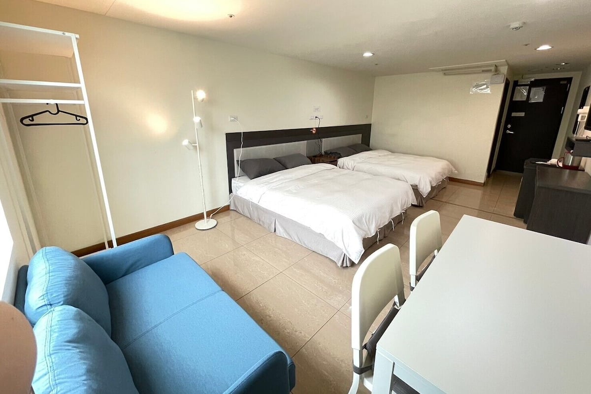 A hotel room with two beds, a desk, a chair, a wardrobe, and a floor lamp.