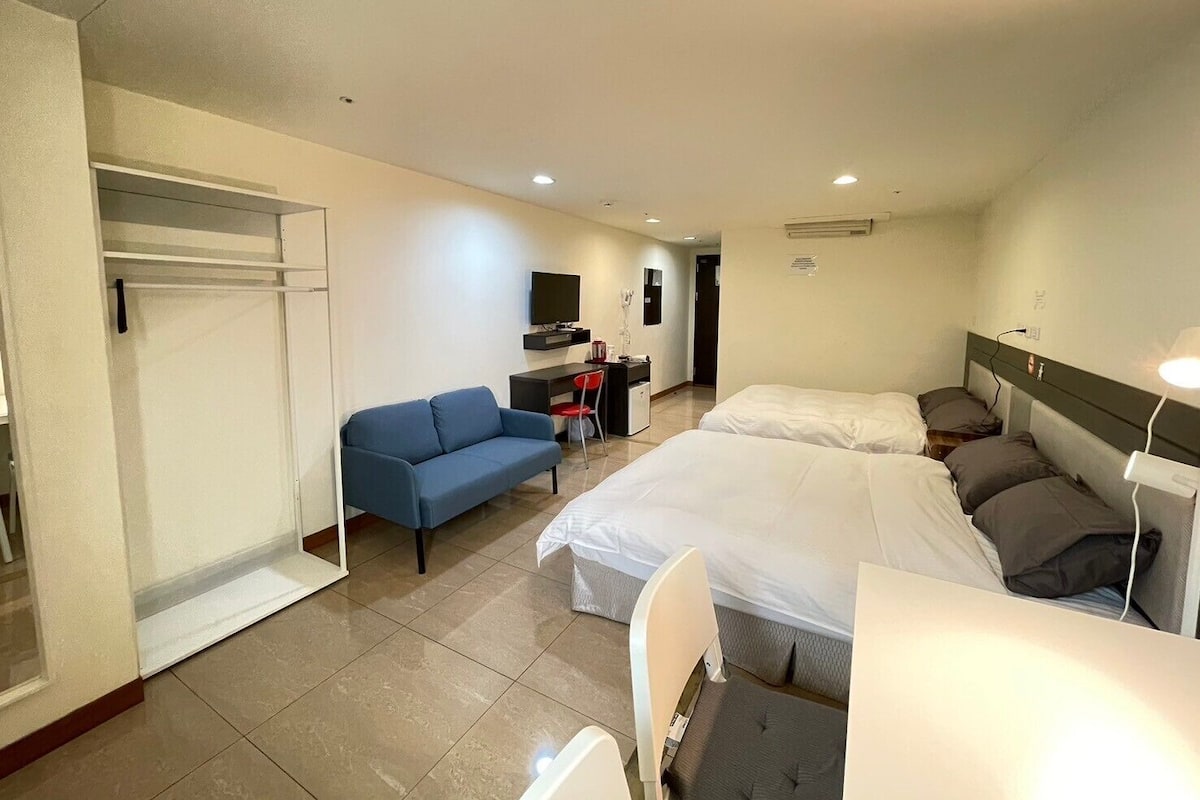A hotel room with two beds, a sofa, a desk with a chair, a TV, and a wardrobe.