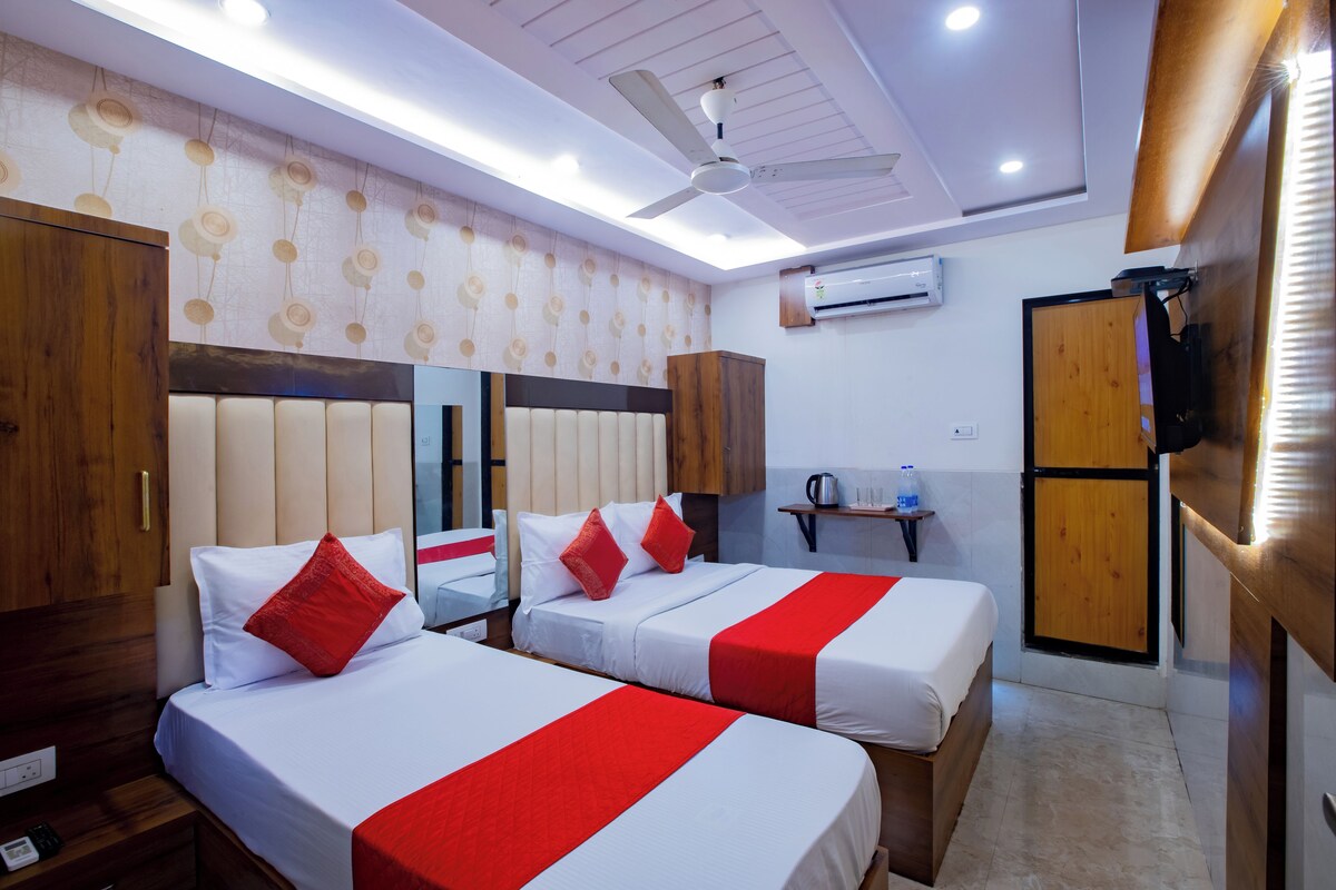 Habitación de hotel con dos camas, ventilador de techo, equipo de aire acondicionado, armario de madera y una pequeña mesa con una tetera y vasos.