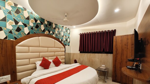 Une chambre moderne avec un grand lit, une tête de lit en bois, un coussin rouge décoratif, une petite table de chevet, un téléviseur et un mur orné d’un motif géométrique.