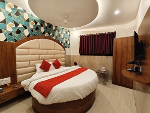 Une chambre moderne avec un grand lit, une tête de lit en bois, un coussin rouge décoratif, une petite table de chevet, un téléviseur et un mur orné d’un motif géométrique.