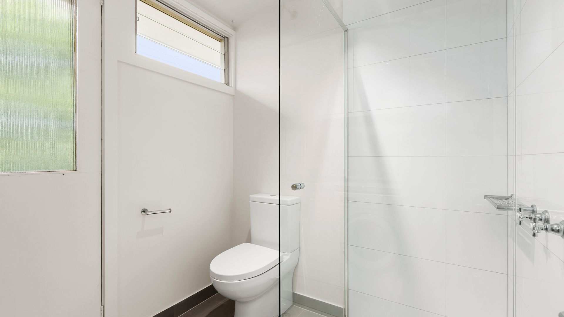 A modern bathroom with a toilet, a glass shower enclosure, and a window above the toilet.