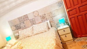 Quarto de hotel com colcha estampada, cabeceira com desenho em mosaico, mesa de cabeceira com abajur e porta vermelha.