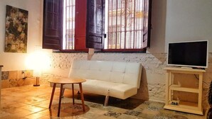 Uma sala de estar com um sofá branco, uma mesinha redonda, uma televisão em um suporte de madeira e uma janela com grades.