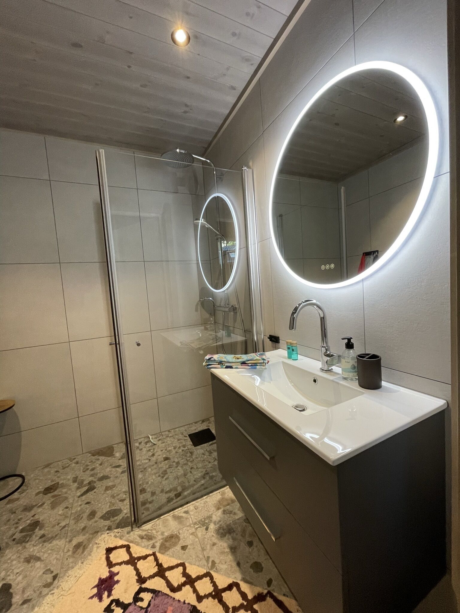 A modern bathroom with a glass shower enclosure, a large mirror with integrated lighting, a white sink, and a dark-toned vanity.