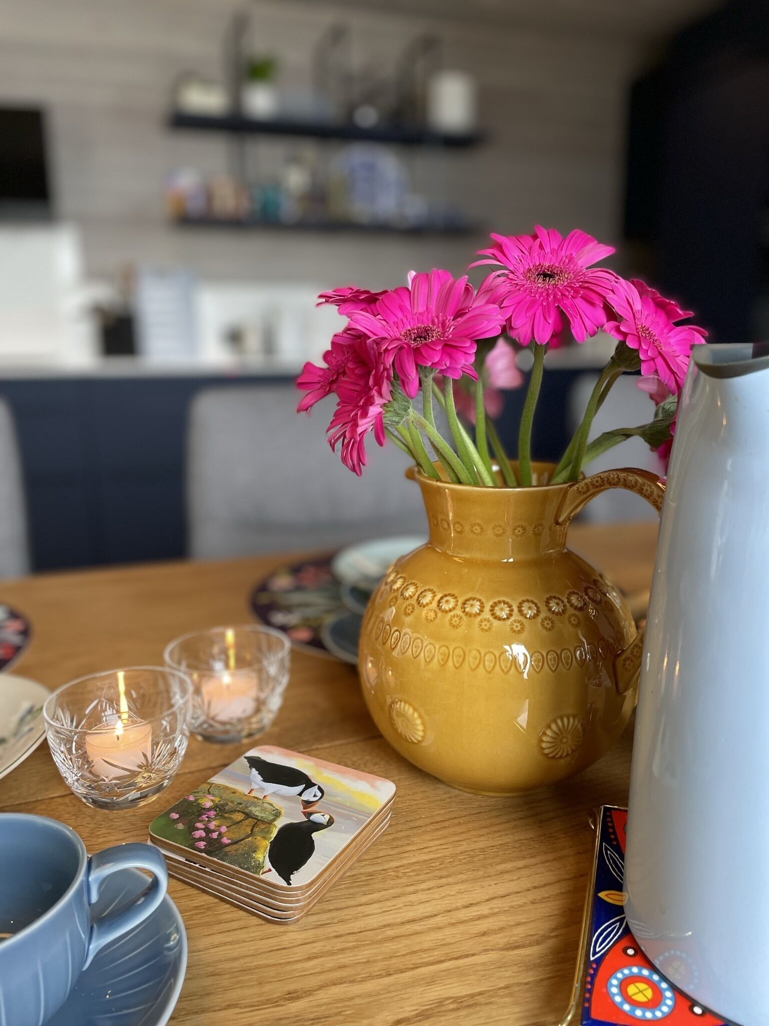 Una mesa con un jarrón amarillo que contiene flores rosas, una pila de cartas y una tetera.