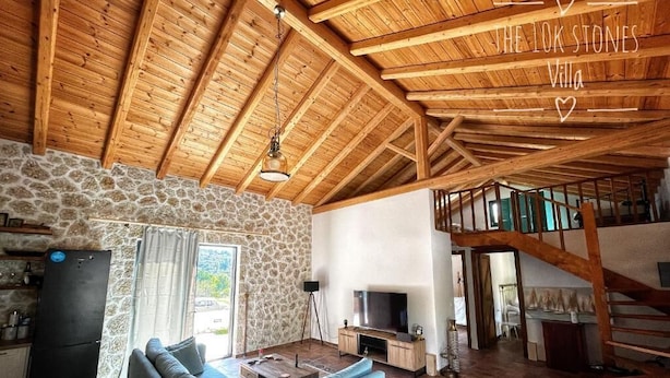 Un interior rústico con pared de piedra, vigas de madera, sofá azul, televisor y escalera.