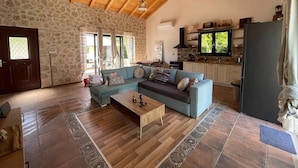 Amplia sala de estar con pared de piedra, techo de madera, un sofá azul, una mesa de centro de madera y una zona de cocina con refrigerador y televisor.