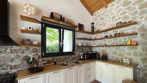 Una cocina con armarios blancos, pared de piedra, estantes de madera y ventana con vistas a follaje.