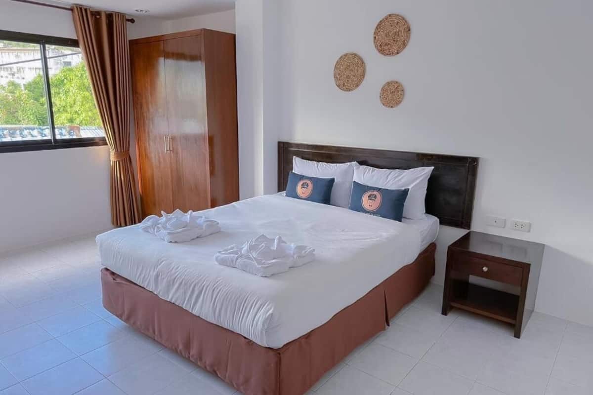 A neatly made bed with white linens and two pillows, a wooden wardrobe, a small bedside table, and a window with curtains.