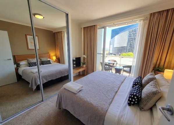 Habitación de hotel con dos camas, un espejo grande, un escritorio, una silla y un balcón con vistas a la ciudad.