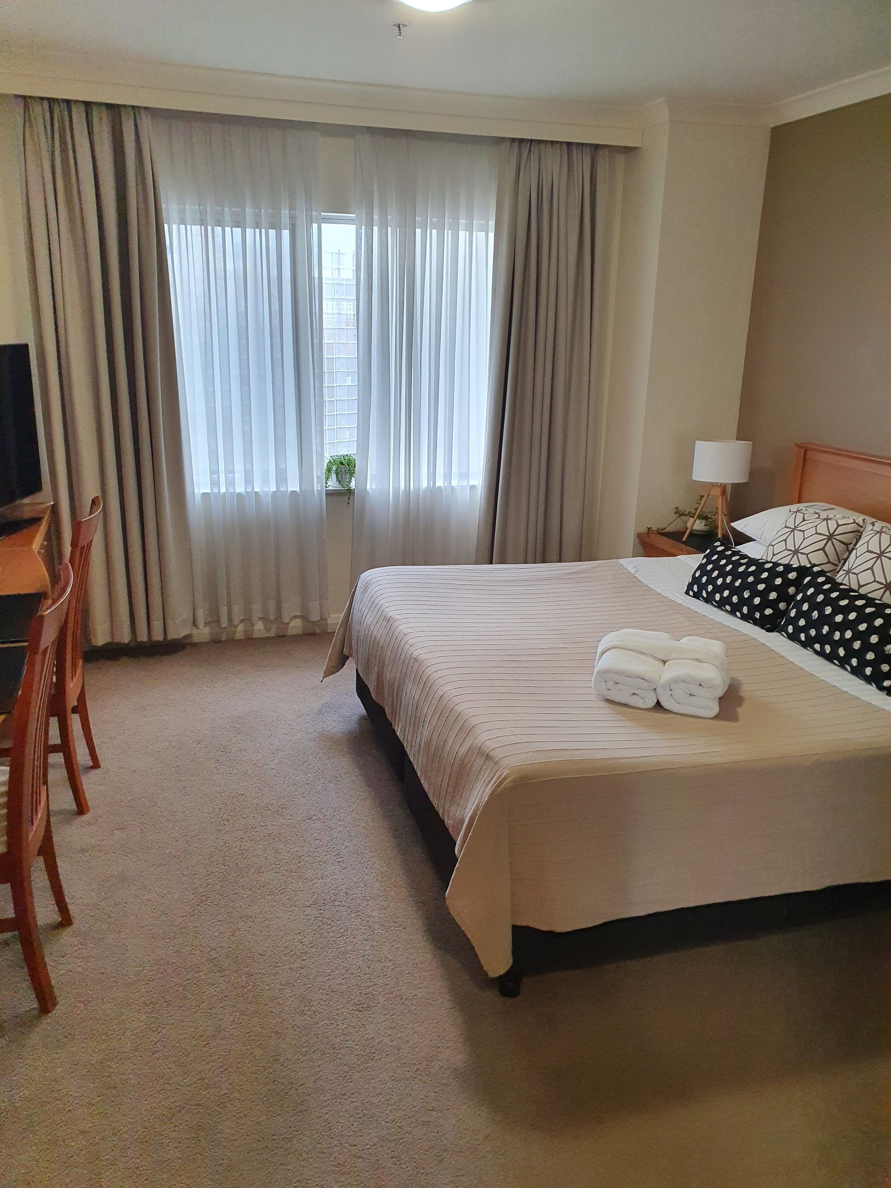 A hotel room with a large bed, a desk with a chair, a nightstand with a lamp, and a window with curtains.