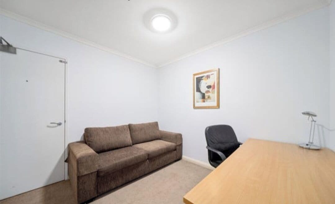 A room with a beige sofa, a wooden table, a black office chair, a framed picture on the wall, and a white door.