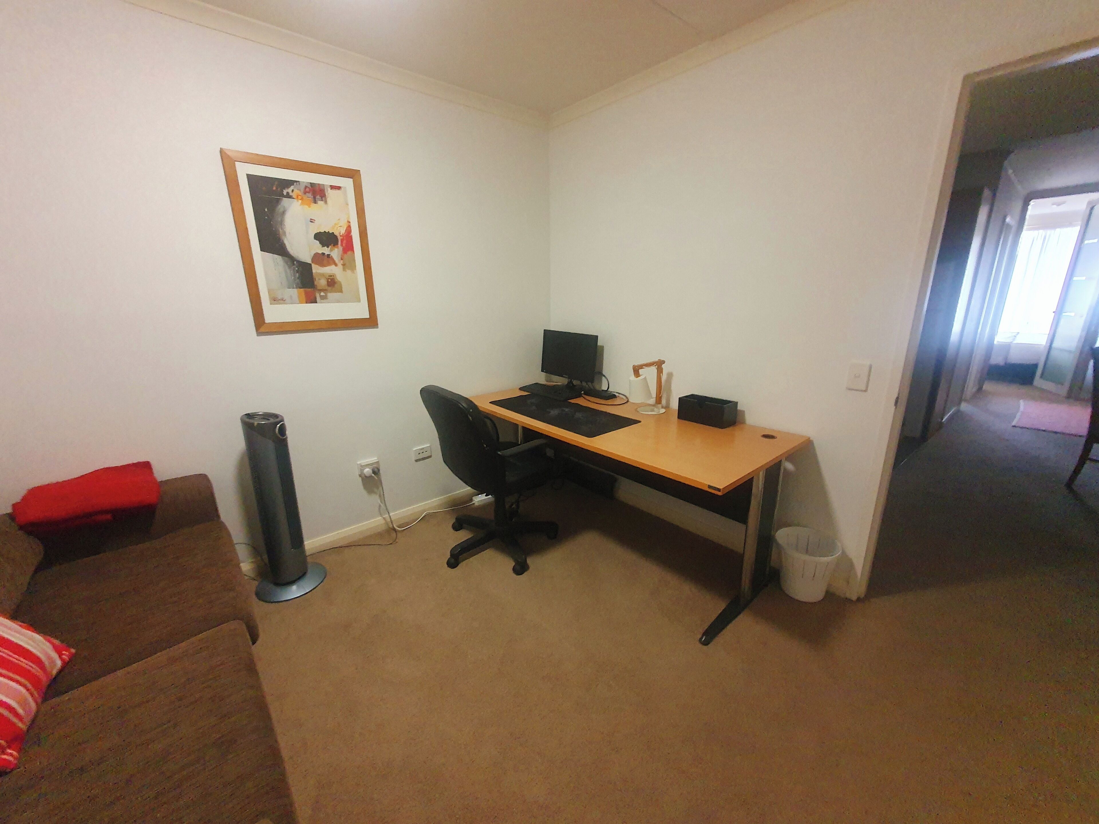 A small office space with a desk, chair, and a computer. There is a couch with a red pillow, a waste bin, and a framed picture on the wall.
