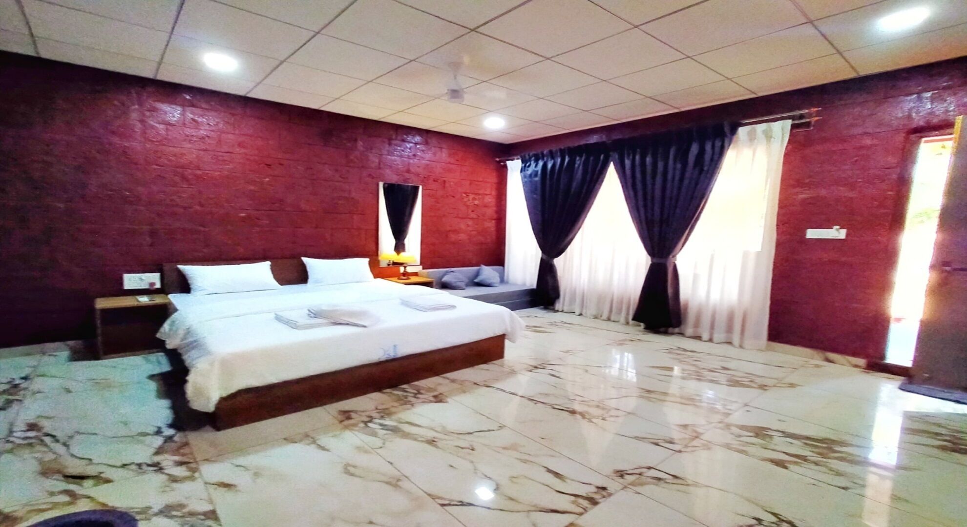 A bedroom with a large bed, a nightstand, a window with curtains, and a red brick wall.
