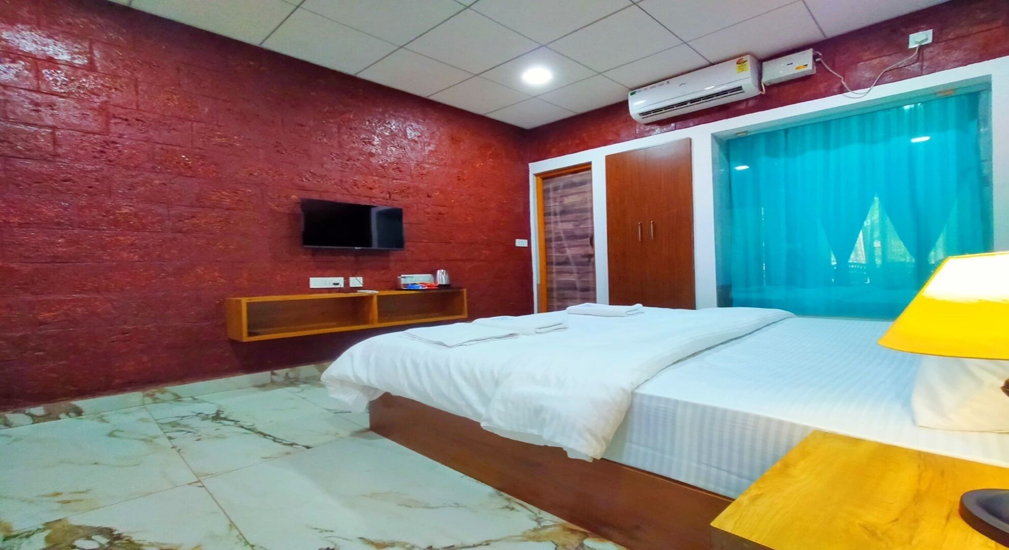 A bedroom with a bed, a TV mounted on the wall, a wooden shelf, and a window with curtains.