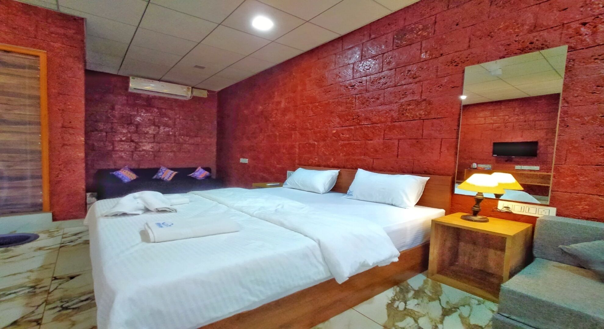 A hotel room with a large bed, a bedside lamp, a TV, and a red brick wall.
