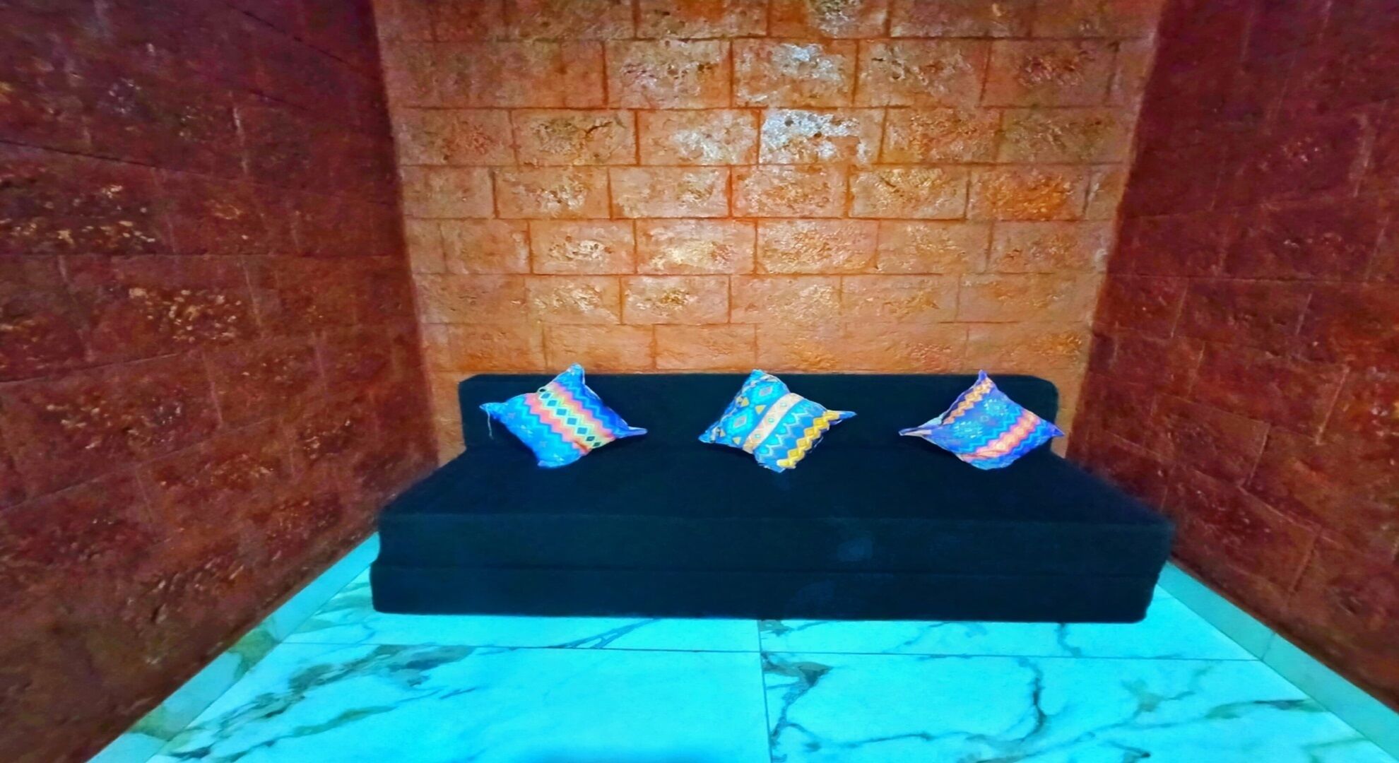 A black sofa with three colorful pillows against a brick wall.