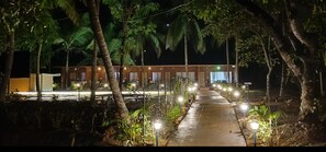 A pathway leading to a building with multiple windows, surrounded by trees and illuminated by street lamps. - West Coast Gokarna (Kumta)