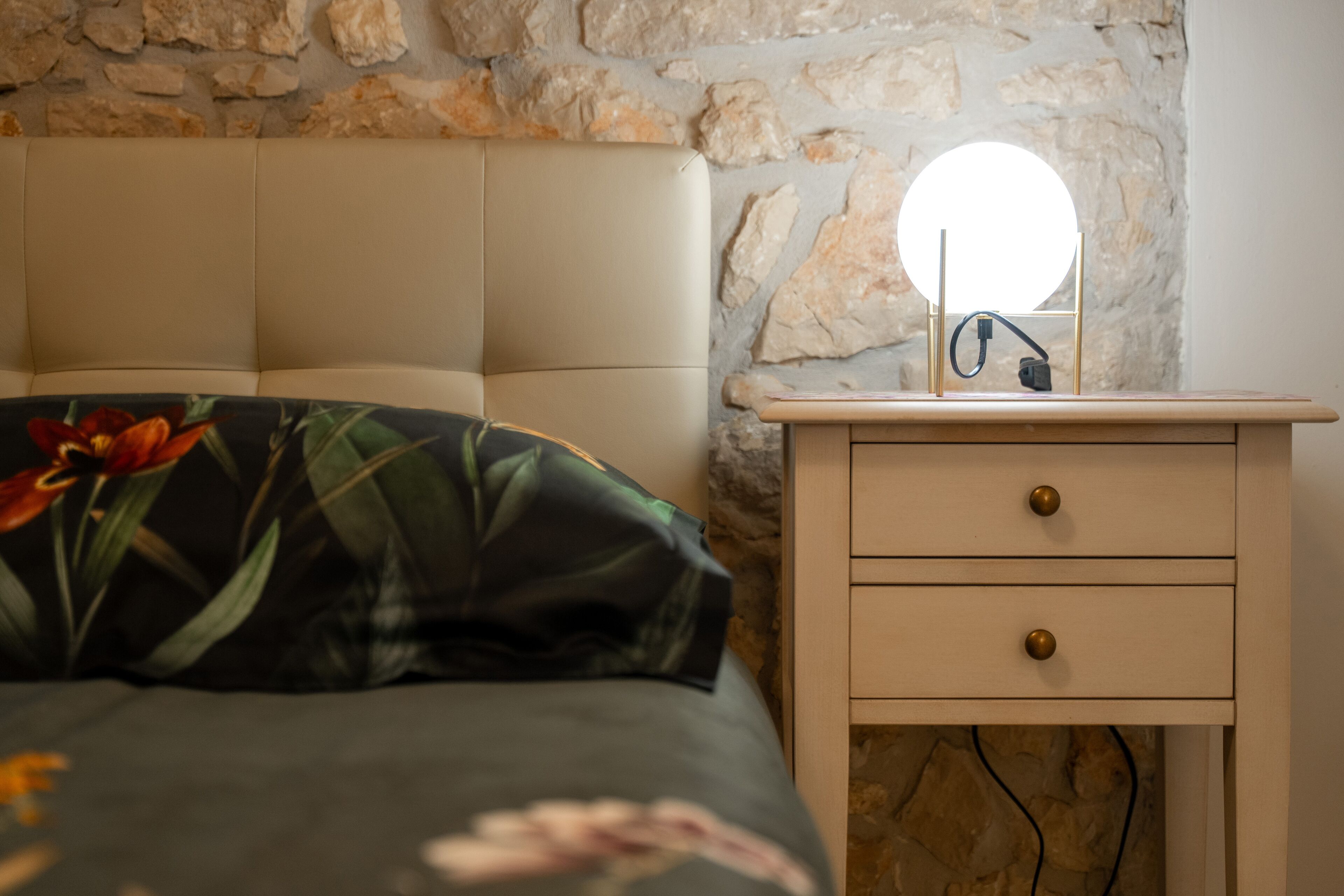 Un lit recouvert d’une couvre-lit à motifs floraux, une table de chevet avec un miroir rond et, en arrière-plan, un mur en pierre.