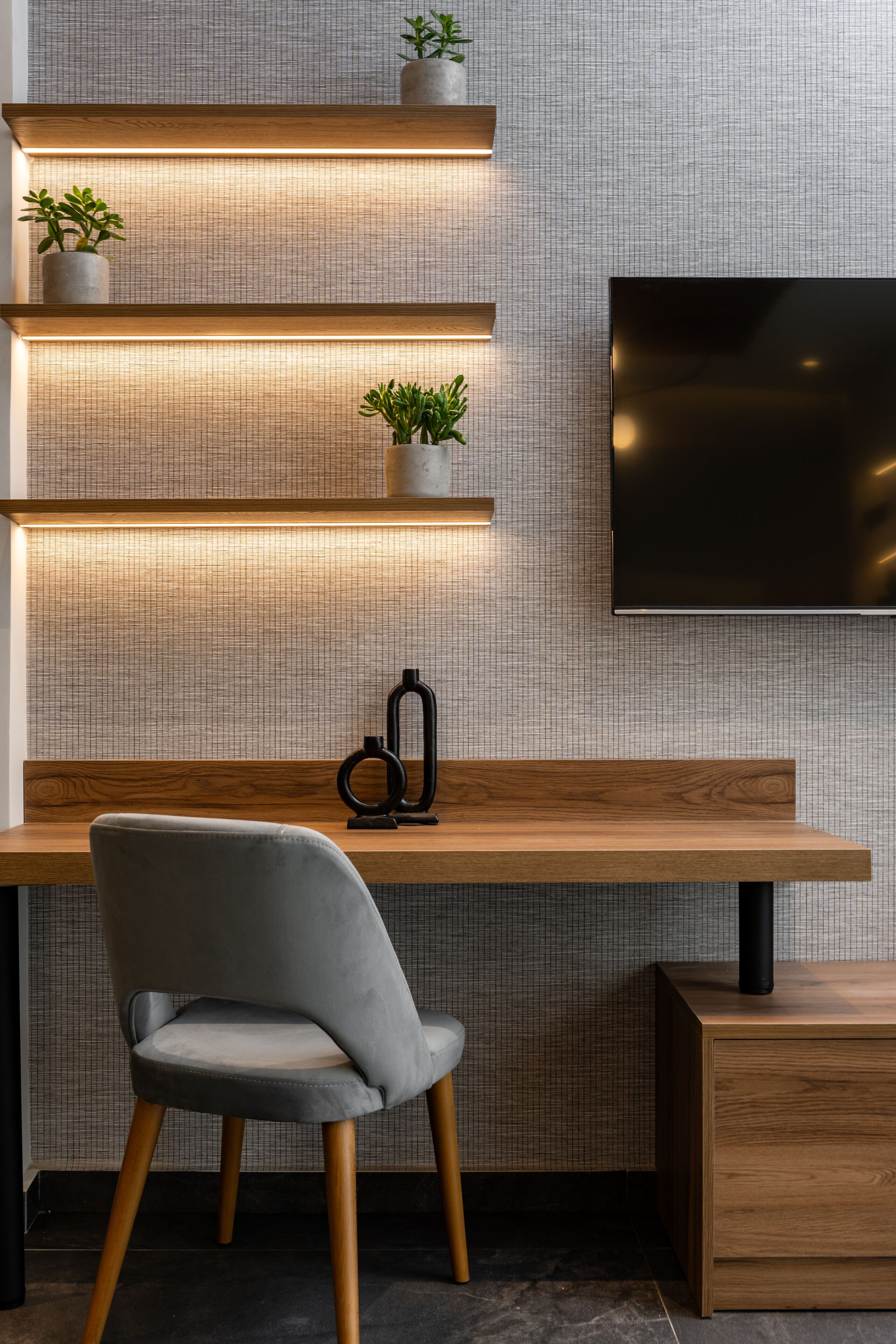 A modern workspace with a wooden desk, a grey upholstered chair, and a mounted TV.