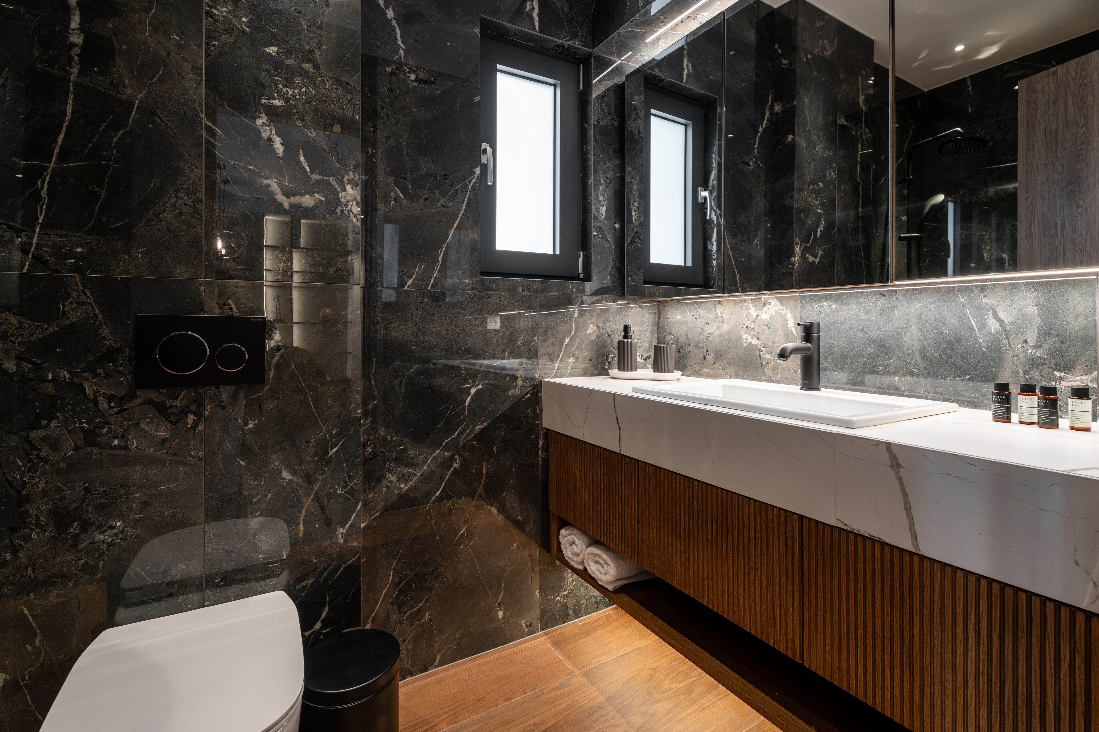 Ein modernes Badezimmer mit Marmorwand, einem großen Spiegel, einem Holzwaschtisch und einer Toilette.