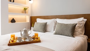 A neatly made bed with white linens, a tray of breakfast including orange juice, pastries, and coffee cups, and a bedside table with a vase and a plant.