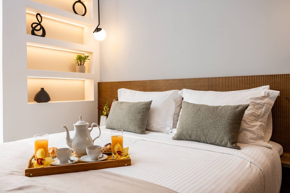 Een netjes opgemaakt bed met witte lakens, een ontbijttafel met jus d'orange, gebak en koffiekopjes, en een nachtkastje met een vaas en een plant.