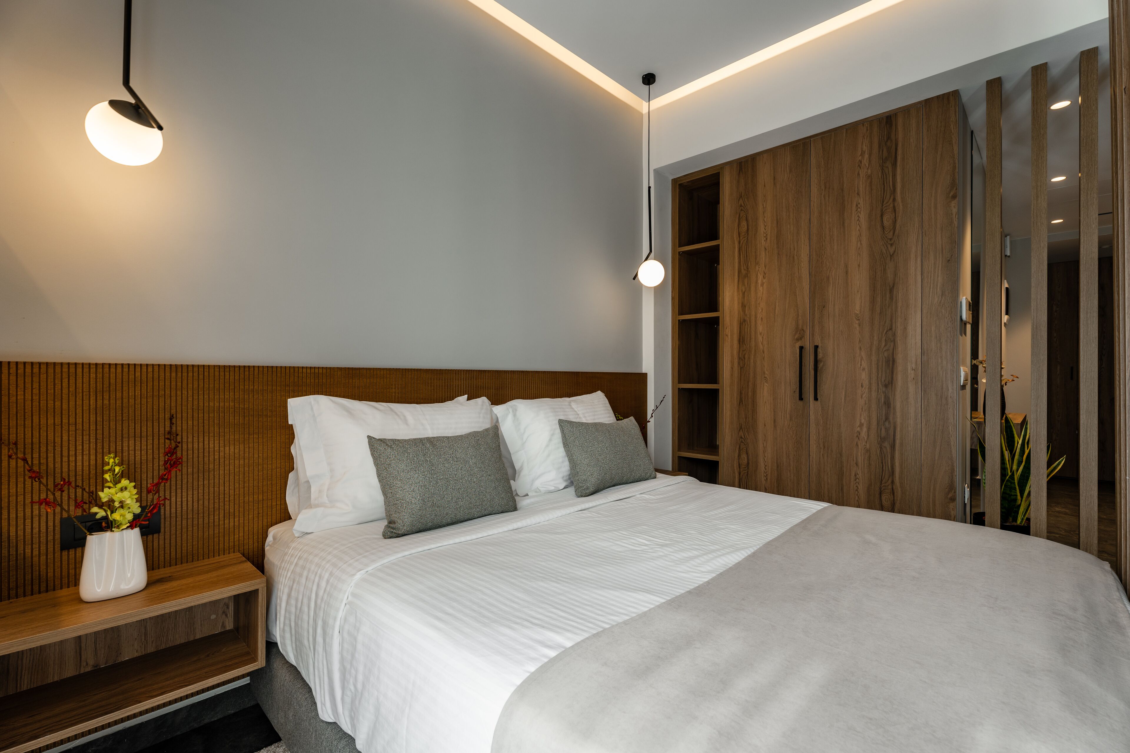 Un dormitorio moderno con cabecera de madera, una cama grande con ropa de cama blanca, una mesita de noche con un jarrón de flores y un armario con espejo.