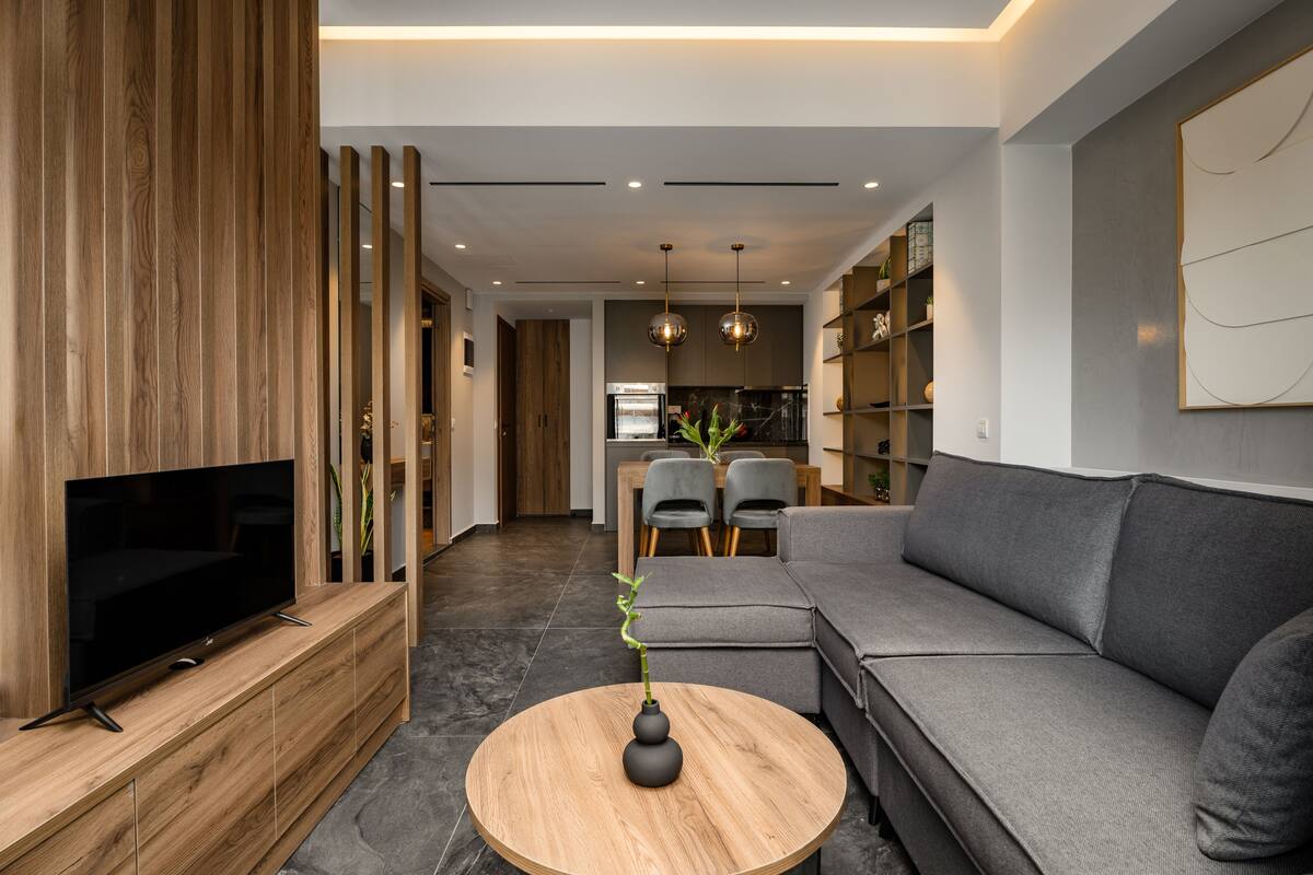 Een moderne woonkamer met een flatscreen televisie, een grijze bank, een houten salontafel en een eethoek met stoelen en een tafel.